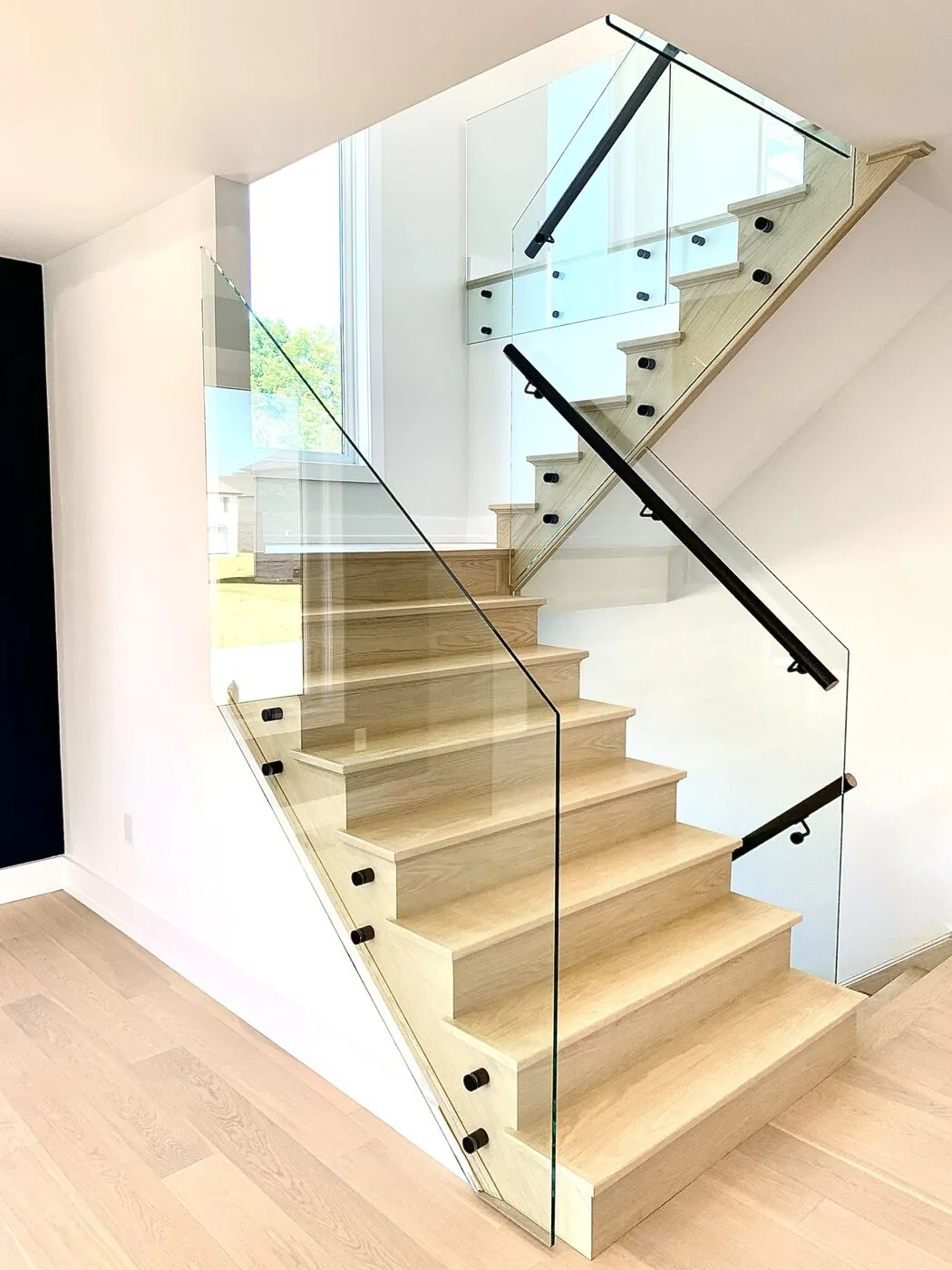 Frameless glass stair railing with black handrail on a light oak staircase, installed by California Glass Railing